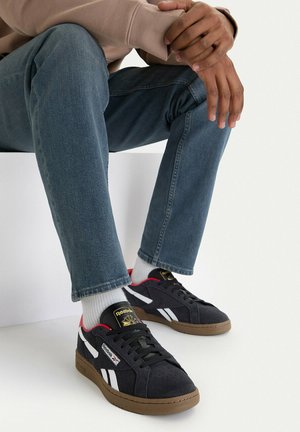 Person trägt dunkelblaue Reebok-Sneaker mit weißen Streifen, weiße Socken, blaue Jeans und einen beigen Pullover und sitzt auf einem weißen Block.