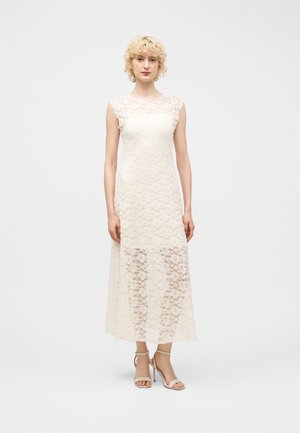 Ung kvinde med kort krøllet blondt hår iført en ærmeløs hvid blonde midi-kjole og beige højhælede sandaler, stående oprejst.