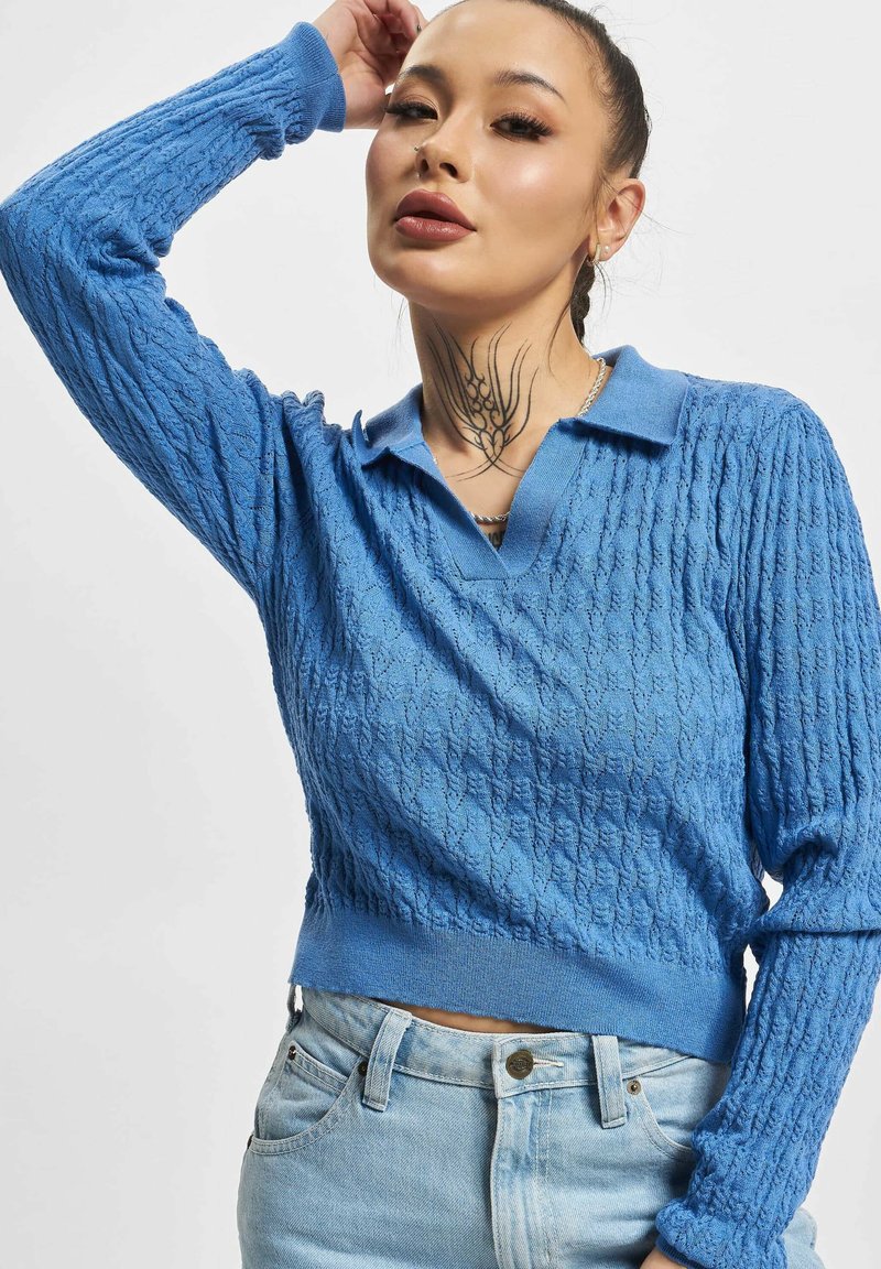 Blå strikket genser med et teksturert mønster, med polo-krage og lange ermer, sammen med lyseblå jeans.
