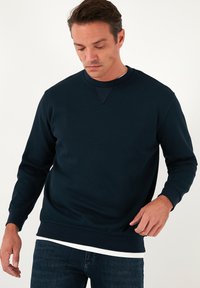 Sötétkék crewneck pulóver struktúrált anyagból, hosszú ujjakkal és diszkrét V-varrással a nyakkivágásnál. Fehér ing fölé viselve.