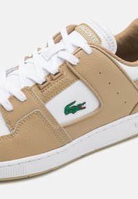Tan ir balti sportbačiai su tekstūruota oda, balti raišteliai ir žalias Lacoste logotipas šone. Ypatybės: lygus guminis padas.