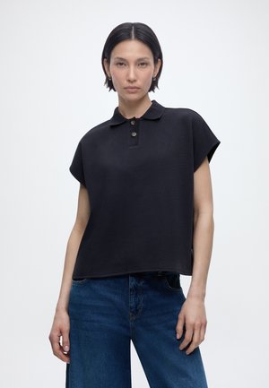 Ung kvinde med kort sort hår iført en løs sort kortærmet polo-shirt og blå denimjeans, stående mod en ensfarvet baggrund.