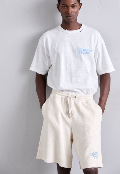 Hvit oversized t-skjorte med blå tekst og slitt utringning, kombinert med teksturerte kremfargede shorts med elastisk midje og sidelommer.