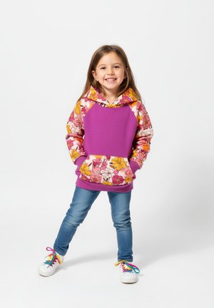 Uśmiechnięta dziewczyna w fioletowej bluzie z kwiatowym wzorem, niebieskich dżinsach i białych trampkach z kolorowymi sznurówkami stoi z rękami w kieszeniach na białym tle.