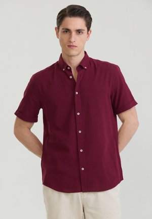 Rövid barna hajú fiatal férfi, aki egy gombolt, rövid ujjú bordó inget és bézs nadrágot visel, a kezeivel a háta mögött áll.