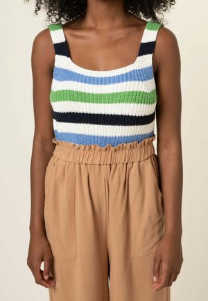 Femme portant un top sans manches en maille rayé bleu, vert, blanc et noir, associé à un pantalon beige taille haute style paperbag.