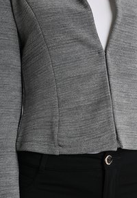 Grauer Blazer mit Struktur, mit Reverskragen und vorderem Reißverschluss. Kombiniert mit schwarzen Hose, die eine runde Metallknopf haben.