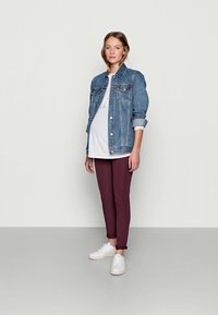 Pregnant woman standing, wearing a white top, blue denim jacket, maroon pants, and white sneakers against a plain light background.