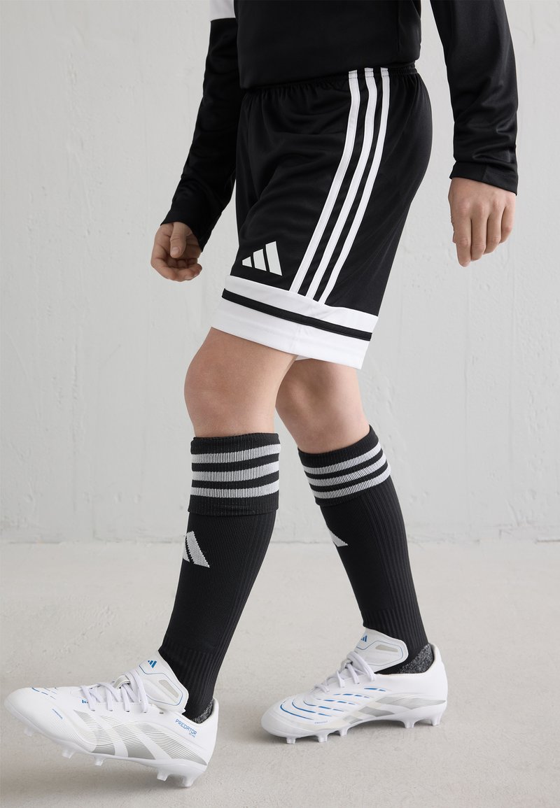Shorts de sport noirs avec des rayures blanches, chaussettes noires jusqu'aux genoux avec des rayures grises, et chaussures de football blanches avec des accents bleus.