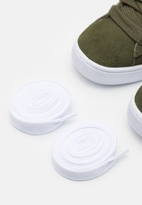 Grøn nubuck-sneaker med hvid gummisål, ledsaget af to ruller hvide snørebånd, tekstureret stof og fladt snørebåndsdesign.