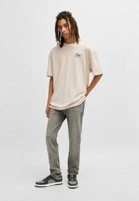 Beżowy, oversize’owy t-shirt z okrągłym dekoltem i czarną naszywką, w połączeniu z szarymi, dopasowanymi dżinsami i czarnymi sneakerami z szarymi akcentami.