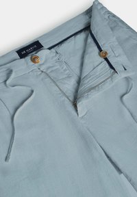Pantalon bleu clair avec fermeture éclair avant partiellement ouverte, bouton marron, taille à cordon et étiquette de la marque Mr Marvis à l'intérieur.