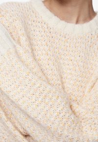 Ljus krämfärgad tröja med en löst stickad design, som har ett strukturerat mönster och guldgula fläckar. Ribbad krage och manschetter.