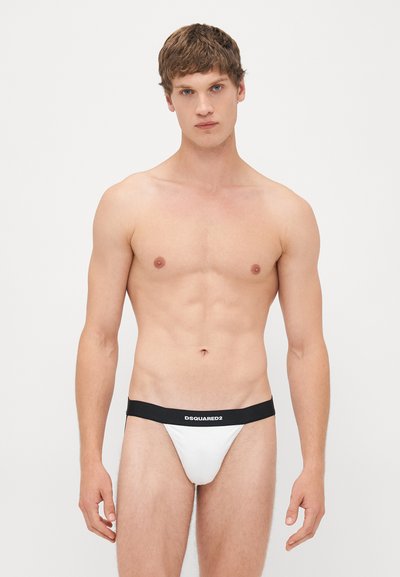Jeune homme aux cheveux bruns courts portant des slips blancs avec une ceinture noire marquée "DSQUARED2" sur un fond blanc uni.