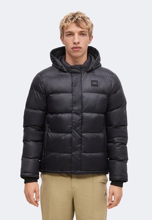 Junger Mann trägt eine schwarze gesteppte Kapuzenjacke und beige Hose und steht vor einem schlichten hellen Hintergrund.