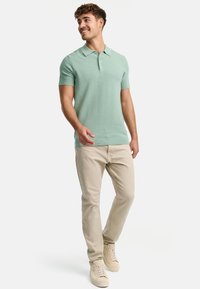 Hellgrünes Poloshirt mit kurzen Ärmeln, geripptem Kragen und strukturiertem Stoff; kombiniert mit beigen Hosen und hellen Sneakers.