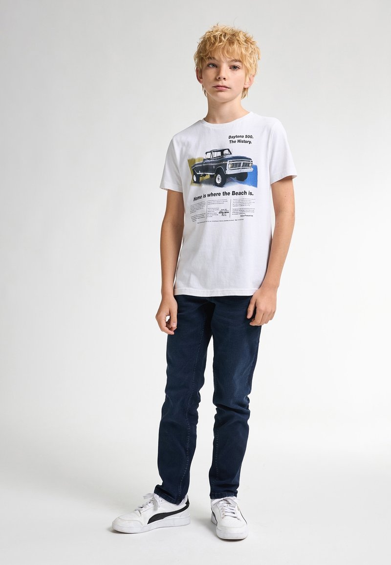 Hvid bomulds T-shirt med trykt grafik af en lastbil, tekst der beskriver Daytona 500s historie, parret med mørkeblå jeans og hvide sneakers.