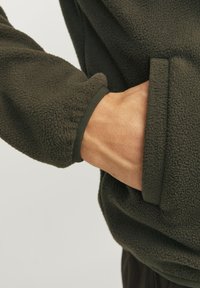 Dark green fleece jacket with a textured surface. The cuff is elasticized, showcasing a snug fit, while a hand is tucked into the side pocket.