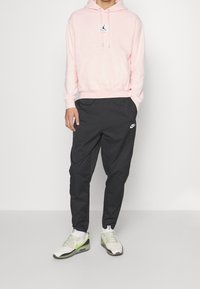 Felpa rosa chiaro con tasca a marsupio e piccolo logo, abbinata a pantaloni neri e scarpe da ginnastica bianche con dettagli verdi e texture in rete.