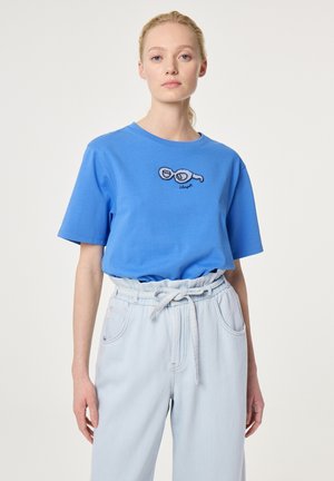 Woman wearing a blue t-shirt with embroidered sunglasses graphic and light blue high-waisted paperbag pants tied at the waist.