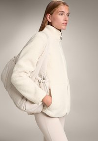 Giacca in pile crema, morbida e soffice, con colletto alto e chiusura con zip, abbinata a una borsa a spalla beige, morbida, con accenti arricciati.