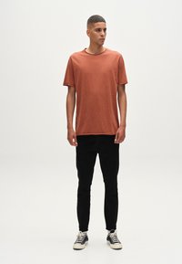 Majica boje hrđe s kratkim rukavima, nosi se uz crne slim-fit hlače i tamnoplave tenisice s bijelim potplatima i vezicama; jednostavna pozadina.