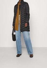Giacca a vento nera con colletto alto, motivo trapuntato e maniche lunghe, indossata sopra un cardigan senape, abbinata a jeans larghi di un azzurro chiaro.