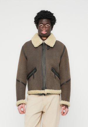 Casaco de shearling castanho com um colarinho e punhos forrados de pelúcia de cor creme. Apresenta um fecho de correr à frente e dois bolsos com fecho de correr em ângulo.