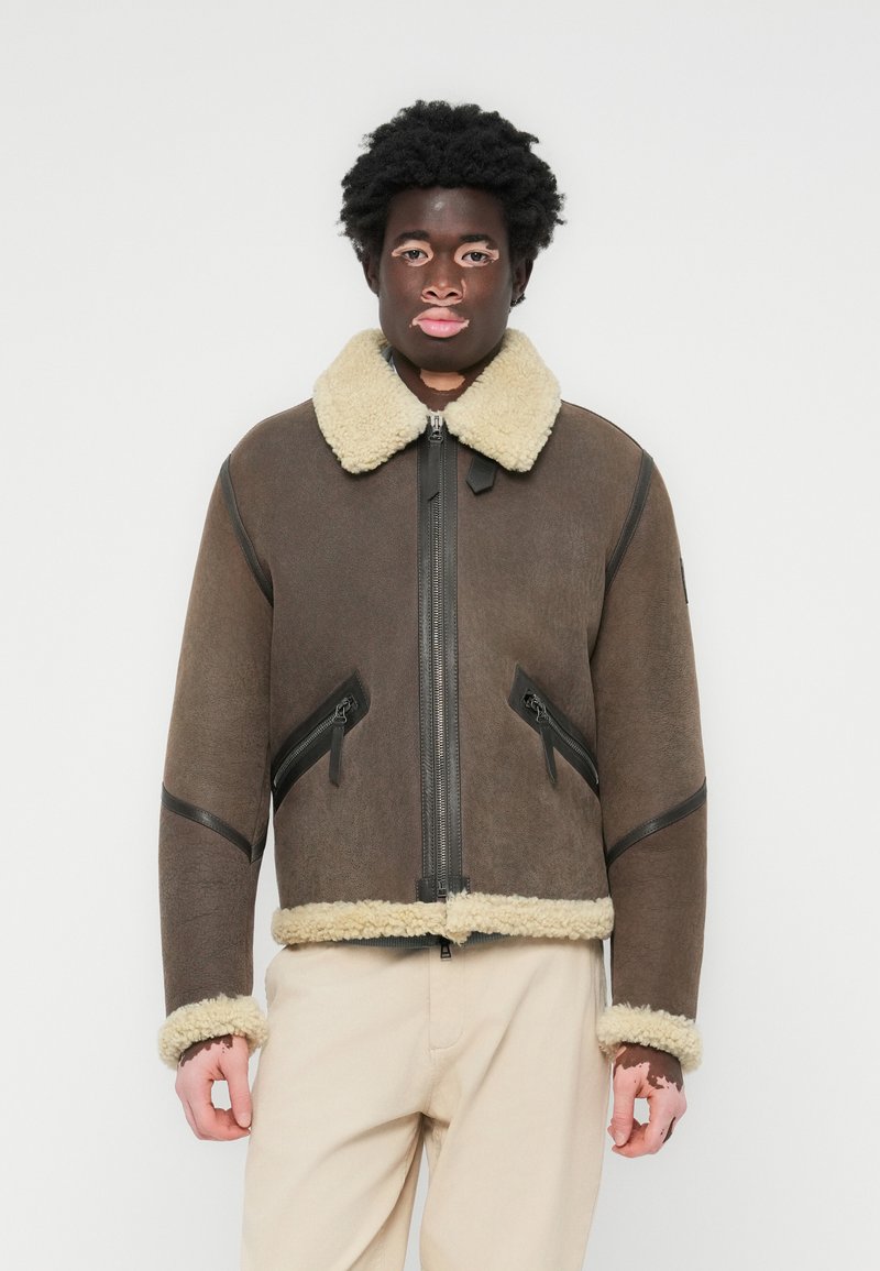 Chaqueta de borreguillo marrón con un cuello y puños forrados de piel color crema. Cuenta con un cierre frontal de cremallera y dos bolsillos con cremallera en ángulo.