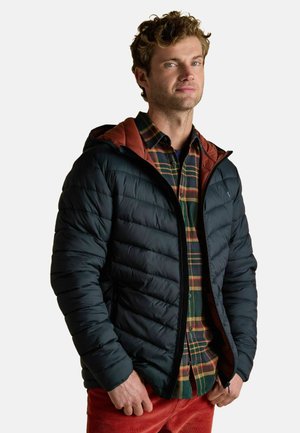 Mann mit lockigem Haar, der eine schwarze Daunenjacke über einem grün-orange karierten Hemd und roten Cordhosen trägt, steht mit den Händen in den Taschen.