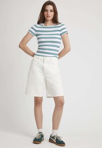 Chemise à manches courtes rayée en blanc, bleu et vert, associée à un short en denim blanc. Le modèle porte des baskets vertes et blanches avec des lacets crème.
