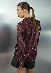 Mujer con una coleta que lleva una blusa de satén color burdeos con lunares blancos y pantalones cortos negros, de pie de lado contra un fondo simple.