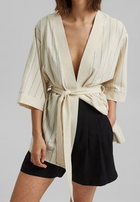 Beige kimonomallinen takki pystysuorilla raidoilla, leveillä hihoilla, syvällä V-aukkolla ja sidotulla vyötärön yksityiskohdalla, yhdistettynä mustiin shortseihin.