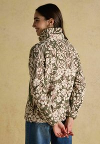 Fleece bunda se květinovým vzorem v krémových a zelených tónech, s vysokým límcem a bočními zipy, nošena s modrými džíny.