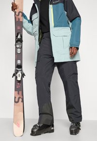 Slidinėjimo įranga apima šviesiai mėlyną ir tamsią striukę, juodas kelnes ir juodus slidinėjimo batus, laikant rudus slides su juodais tvirtinimais ir raudonais akcentais.