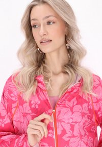 Giacca rosa con zip, caratterizzata da un motivo floreale, tessuto leggero e zip arancione a contrasto. Il modello tiene la cerniera, mettendo in evidenza i dettagli del design.