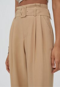 Pantalons taille haute beige à jambe large, avec une ceinture à boucle carrée, des plis sur le devant et des poches latérales. Texture lisse.