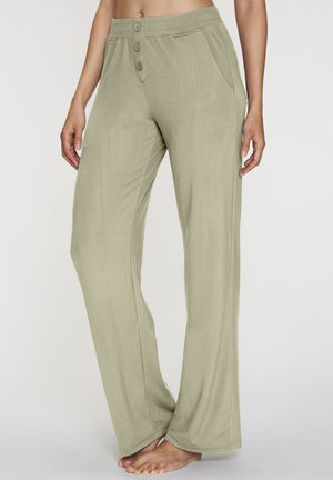 Woman wearing loose, light olive green pants with two buttons and side pockets, standing barefoot on a plain background.