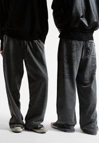 Γκρι παντελόνια φόρμας με χαλαρή εφαρμογή, διαθέτουν κορδόνι στη μέση και κεντημένα σχέδια στο δεξί πόδι, συνδυασμένα με μαύρα αθλητικά παπούτσια.