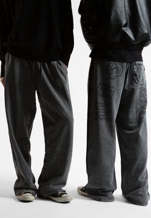 Grå tygbyxor i en avslappnad passform, med dragsko i midjan och broderade detaljer på höger ben, tillsammans med svarta sneakers.