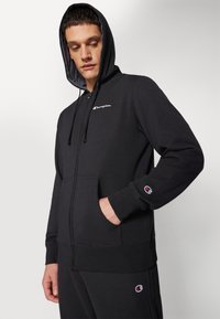 Sudadera con cremallera negra hecha de una mezcla de algodón, con un bolsillo frontal, capucha ajustable con cordón y logo de Champion bordado en el pecho.