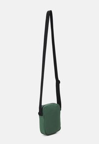 Borsa a tracolla verde realizzata in tessuto morbido con forma rettangolare, caratterizzata da una tracolla nera regolabile e una chiusura con zip sulla parte superiore.