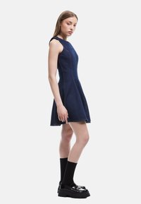 Marineblauwe denim fit-and-flare jurk met mouwloos ontwerp, gestructureerde stof en contrasterende stiksels. Gemaakt met zwarte kniehoge sokken en plateauschoenen.