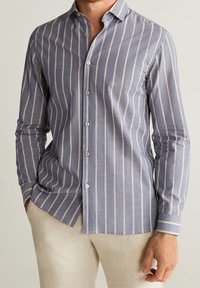 Homme portant une chemise à manches longues bleue avec des rayures verticales blanches, un pantalon beige, une main dans la poche et l'autre détendue le long du corps.