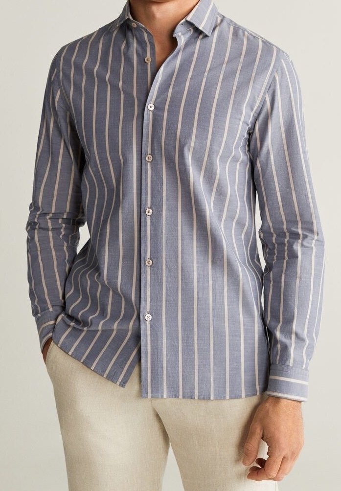 Homme portant une chemise à manches longues bleue avec des rayures verticales blanches, un pantalon beige, une main dans la poche et l'autre détendue le long du corps.