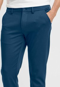 Moški nosi temno modre formalne hlače s sprednjimi žepi, zankami za pas in belim srajčnim delom, roka je v desnem žepu.