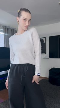 Suéter de malha cinza claro, de mangas longas e com gola redonda, combinado com calças pretas de cintura com cordão, apresentando um corte descontraído.