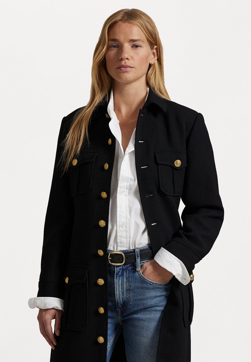 Manteau en laine noir avec des boutons dorés, poches poitrine doubles et coupe droite. Porté sur une chemise blanche et un jean bleu, avec des manches retroussées.