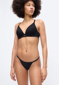 Γυναίκα με σγουρά μαλλιά, φοράει μαύρο αθλητικό σουτιέν και αντίστοιχο στρινγκ, στέκεται μπροστά σε απλό ανοιχτό φόντο.