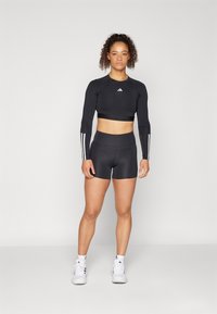 Cropped top sportivo nero con maniche lunghe e tre strisce bianche su ogni braccio, abbinato a pantaloni corti neri a vita alta e sneakers bianche.